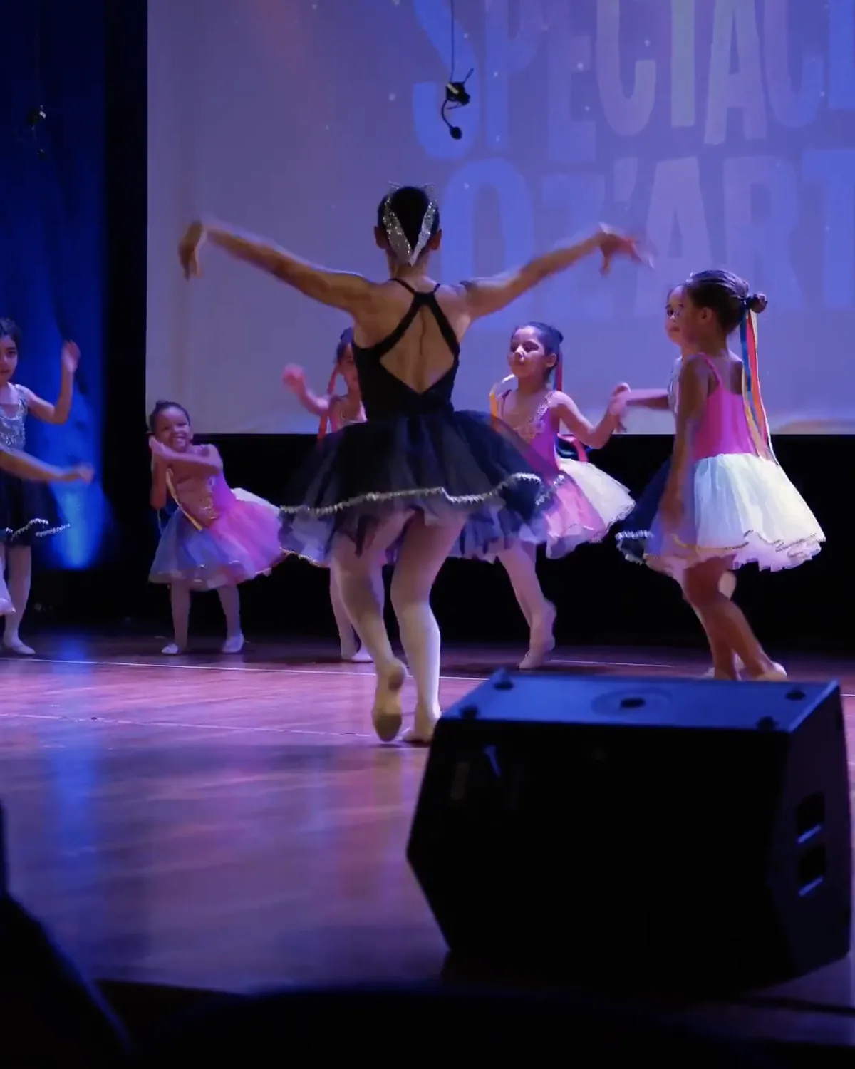 Un groupe de jeunes danseuses en tutu coloré évolue sur scène avec leur professeure.